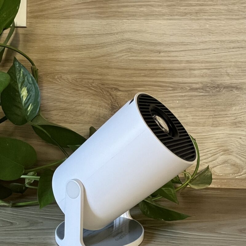 Accueil Projecteur d’ambiance Planète Lumi blanc posé sur une surface en bois clair, entouré de feuilles vertes, illustrant un design minimaliste et naturel.